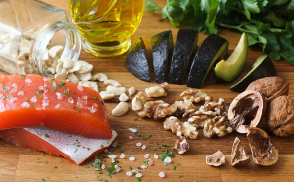 A wooden board with a raw salmon fillet, walnuts, cashews, avocado slices, olive oil, fresh herbs, and sea salt.