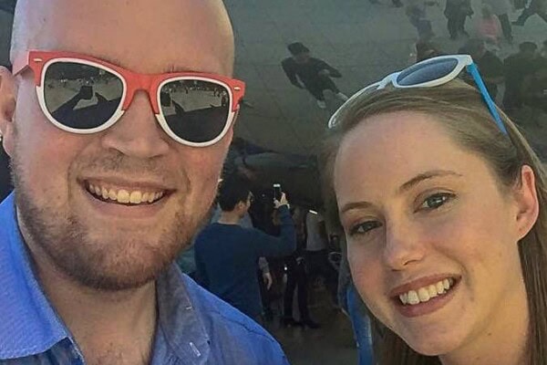 Two people wearing sunglasses smile at the camera, with a reflective surface and several people visible in the background.