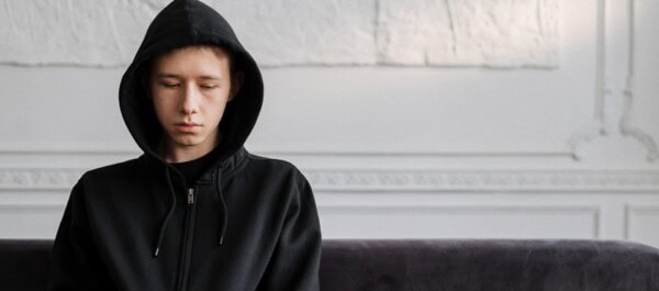 A young person wearing a black hoodie sits on a dark couch, looking down with a neutral expression in a bright, minimalist room.