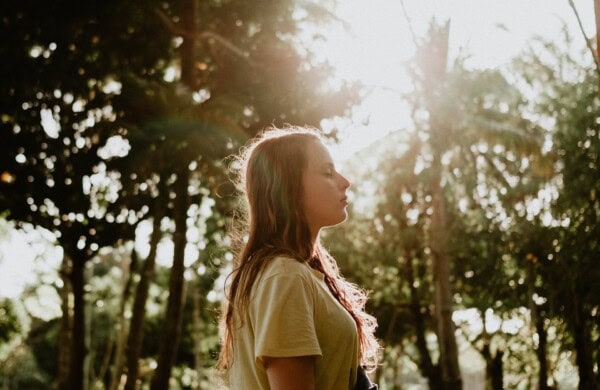 A person with long hair stands outdoors in a wooded area with eyes closed, facing sunlight filtering through the trees.