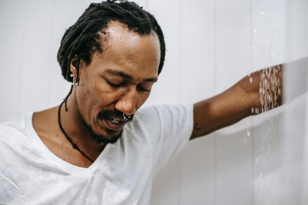 A man with closed eyes stands under running water in a shower, wearing a white t-shirt. His arm is raised, and water droplets are visible on his face and shirt.