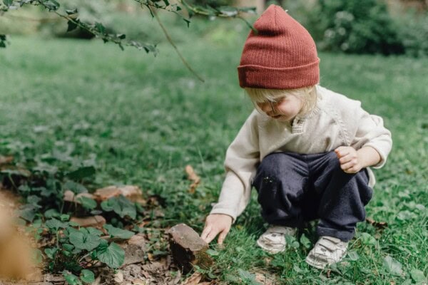 A young child wearing a red beanie squats on the grass and touches a rock in a garden or park setting.