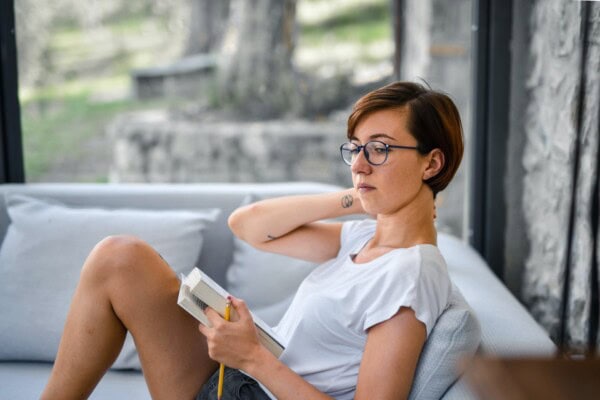 A person with short hair and glasses sits on a sofa, holding an open book and looking thoughtfully to the side.
