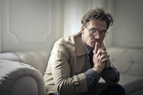 A middle-aged man wearing glasses and a beige coat sits on a sofa, looking thoughtful with his hands clasped near his face.