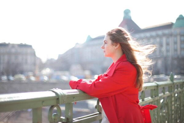 A woman in a red coat stands at a railing, looking into the distance with wind blowing her hair, city buildings blurred in the background.