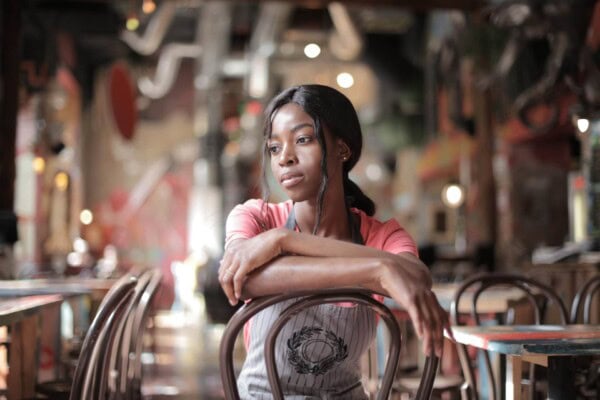 A woman wearing an apron sits with her arms resting on the back of a chair in an empty, dimly lit café, looking thoughtfully into the distance.