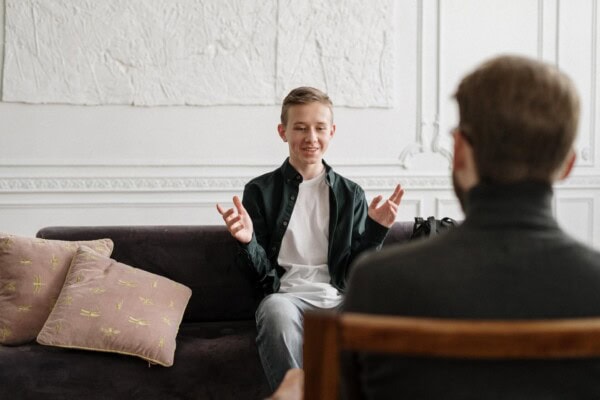 A teenage boy sits on a sofa talking and gesturing with his hands while an adult listens, both in a bright, modern room.
