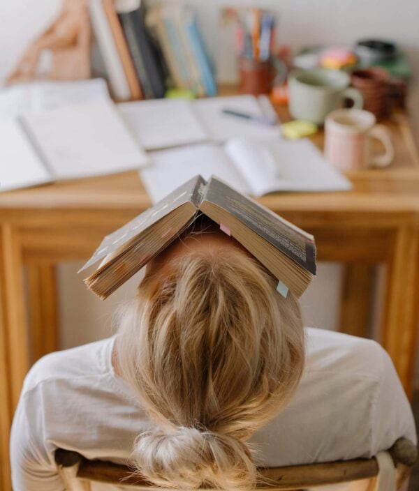 Person with blond hair sits facing away from the camera, with an open book resting on their head, in front of a cluttered desk with papers, books, and mugs.