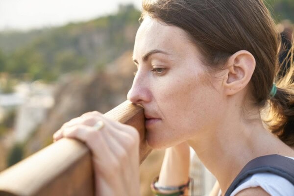 A woman with brown hair tied back rests her chin on a wooden railing, gazing thoughtfully into the distance outdoors.