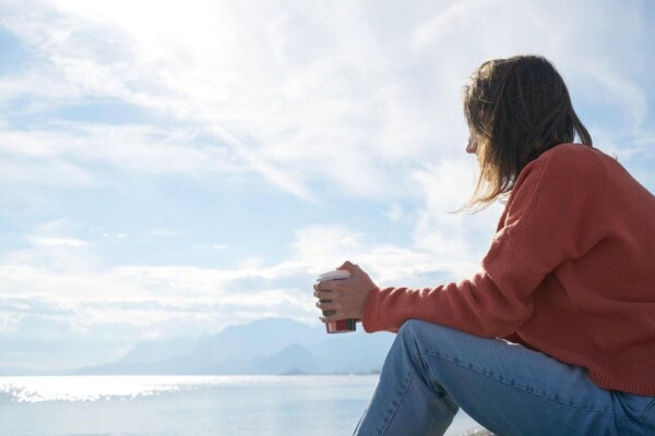 Person in an orange sweater and jeans sits outdoors holding a cup, looking at the sea and mountains under a partly cloudy sky.