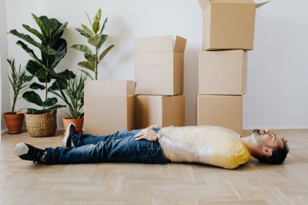 A man wrapped in plastic lies on the floor next to several stacked cardboard boxes and potted plants in a room.