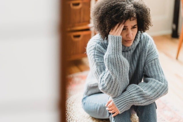 A person with curly hair wearing a gray sweater sits on the floor, resting their head on one hand and appearing distressed or deep in thought.