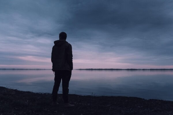 A person stands on the shore looking out over a calm lake at dusk with a cloudy sky and low light.