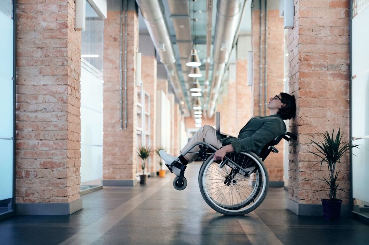 Person in a wheelchair leans back and rests against a brick wall in a modern hallway with plants and exposed ducts.