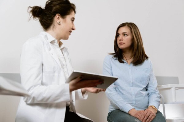 A healthcare professional holding a tablet talks to a seated woman in a medical office.