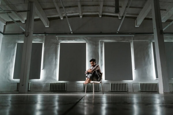 A man sits alone on a chair in the center of a large, empty room with high ceilings and covered windows, hugging his knees and looking down.