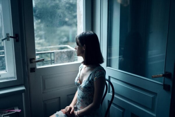 A woman in a floral dress sits on a chair indoors, looking out of a window with soft natural light illuminating her profile.