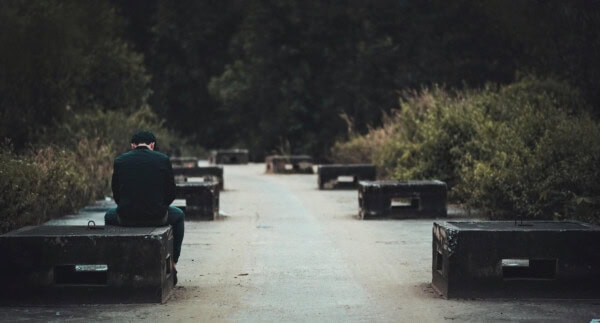 A person sits alone on a concrete bench along a deserted pathway surrounded by greenery, facing away from the camera.