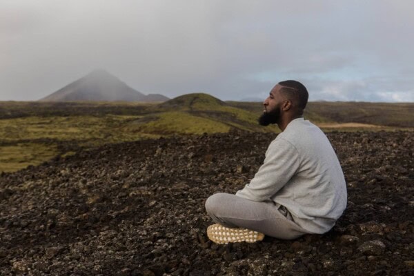 A man sits cross-legged on rocky ground, meditating outdoors with hills and a misty mountain in the background.