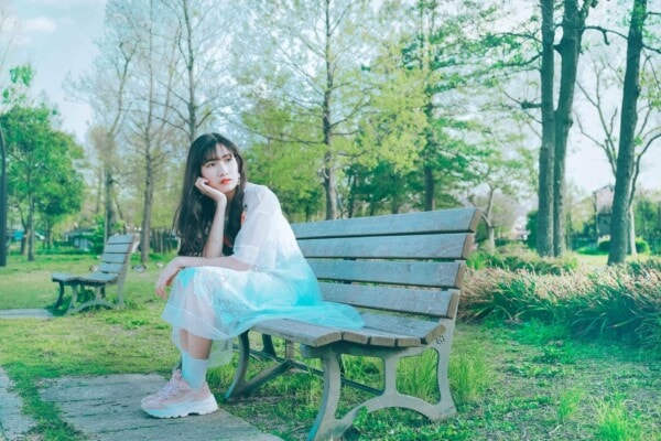 A young woman in a light blue dress sits on a wooden park bench, resting her chin on her hand, surrounded by green trees and grass.