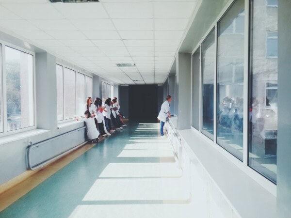 A group of people in white coats stand and talk by the windows in a bright hallway, while one person stands alone facing another window.