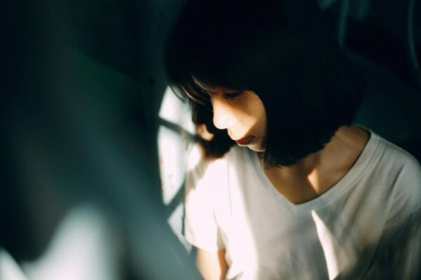 A woman with shoulder-length dark hair and a white shirt looks down, partially illuminated by sunlight and shadow.