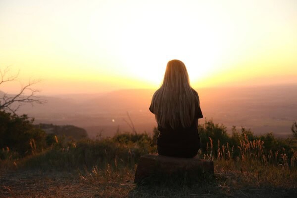 A person with long hair sits on a rock facing the sunset, overlooking a landscape with grass and distant hills.