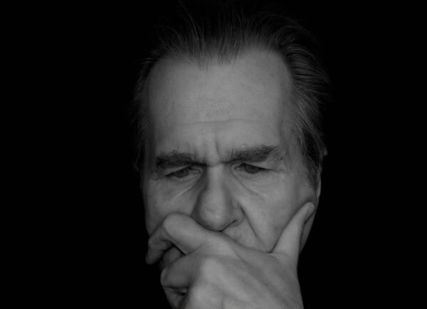 An older man with gray hair rests his chin on his hand, appearing deep in thought against a black background.
