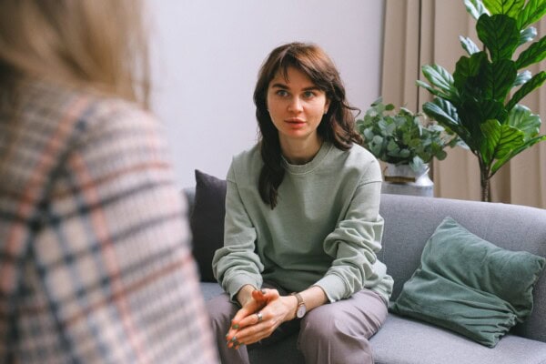 A woman sits on a gray sofa, leaning forward and listening attentively to another person whose back is visible in the foreground. There are green plants and cushions in the background.