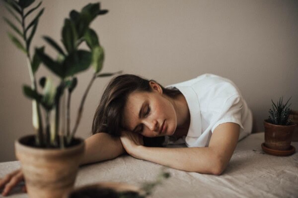 A woman in a white shirt rests her head on her arms on a table with potted plants in the foreground and background.