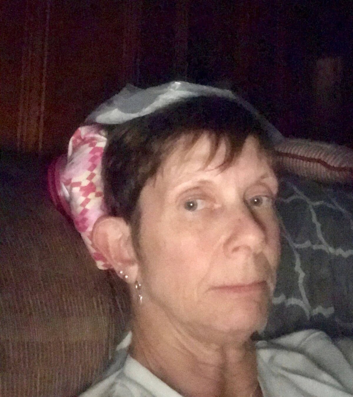 A woman sits indoors with a patterned ice pack resting on her head, looking at the camera with a neutral expression.