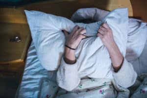 Person lying in bed covering their face with a pillow, with both hands gripping the pillow tightly.