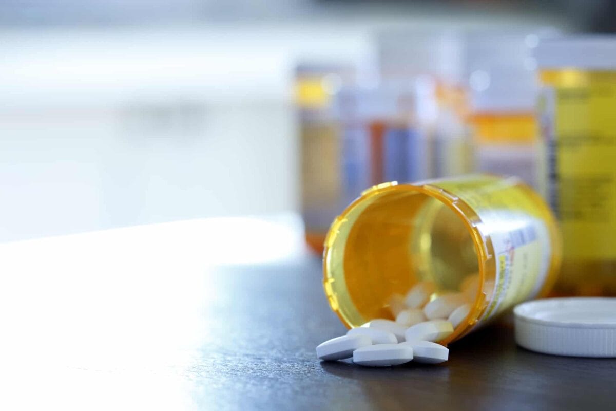 A prescription pill bottle tipped over on a table with white tablets spilling out, and other medicine bottles blurred in the background.