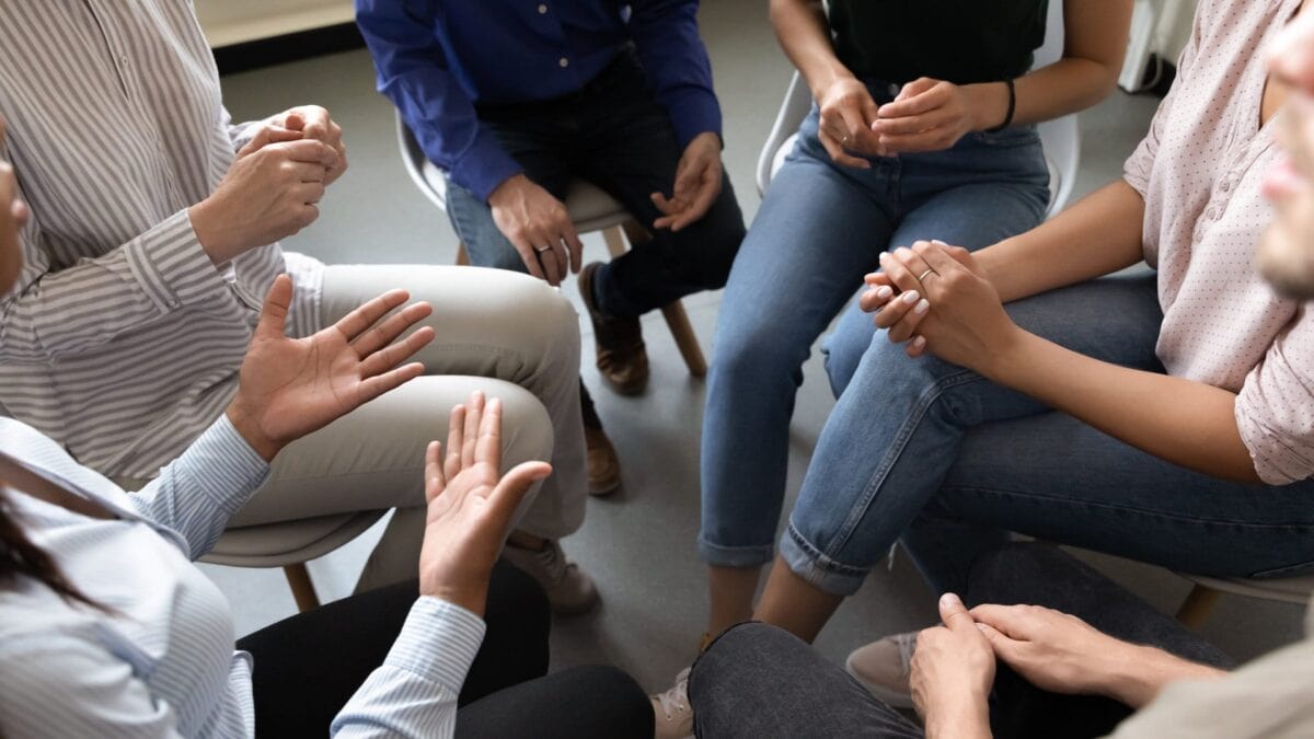 A group of people sit in a circle on chairs, engaged in discussion, with only their hands and laps visible.