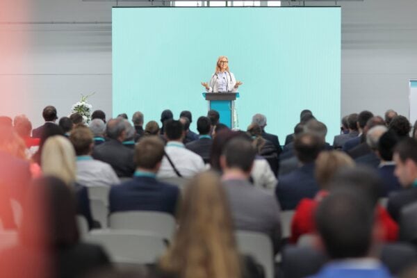 A woman stands at a podium speaking to an audience seated in rows, with a large blank screen behind her in a bright conference hall.