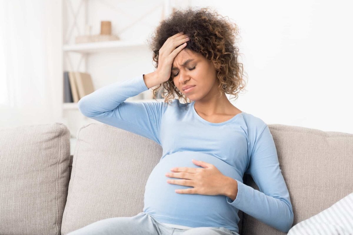 Pregnant woman sitting on a couch, holding her stomach with one hand and her forehead with the other, appearing to be in discomfort.