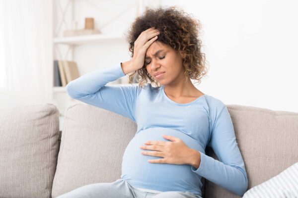 Pregnant woman sitting on a couch, holding her stomach with one hand and her forehead with the other, appearing to be in discomfort.