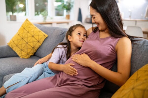 A young girl smiles and rests her hand on the belly of her pregnant mother as they sit together on a gray couch in a living room.