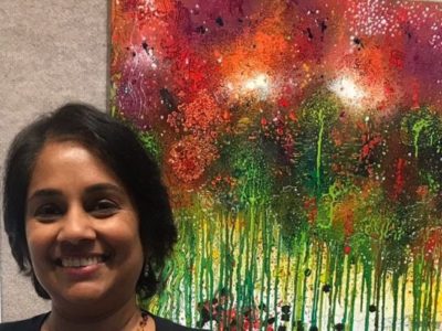 A woman smiles in front of a colorful abstract painting featuring red, orange, and green tones with splattered and dripping paint effects.