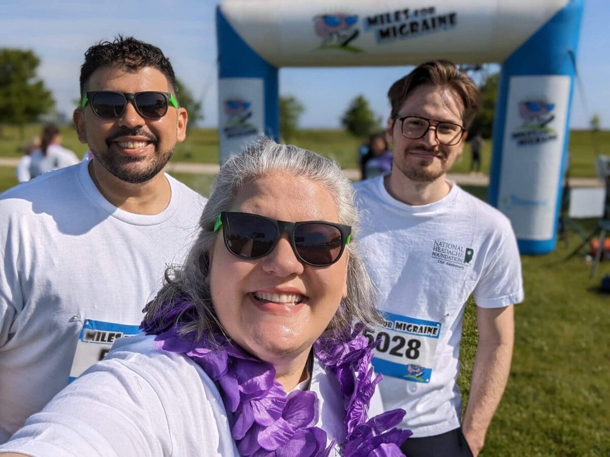 Three people wearing white shirts and sunglasses pose for a photo at an outdoor charity run event, standing in front of an inflatable "Miles for Migraine" arch.