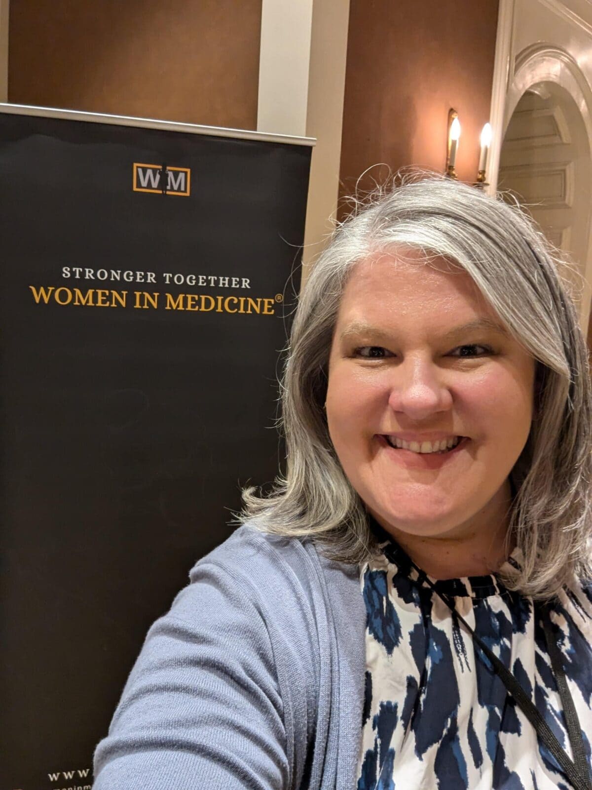 A woman smiling and taking a selfie in front of a "Women in Medicine" event banner indoors.