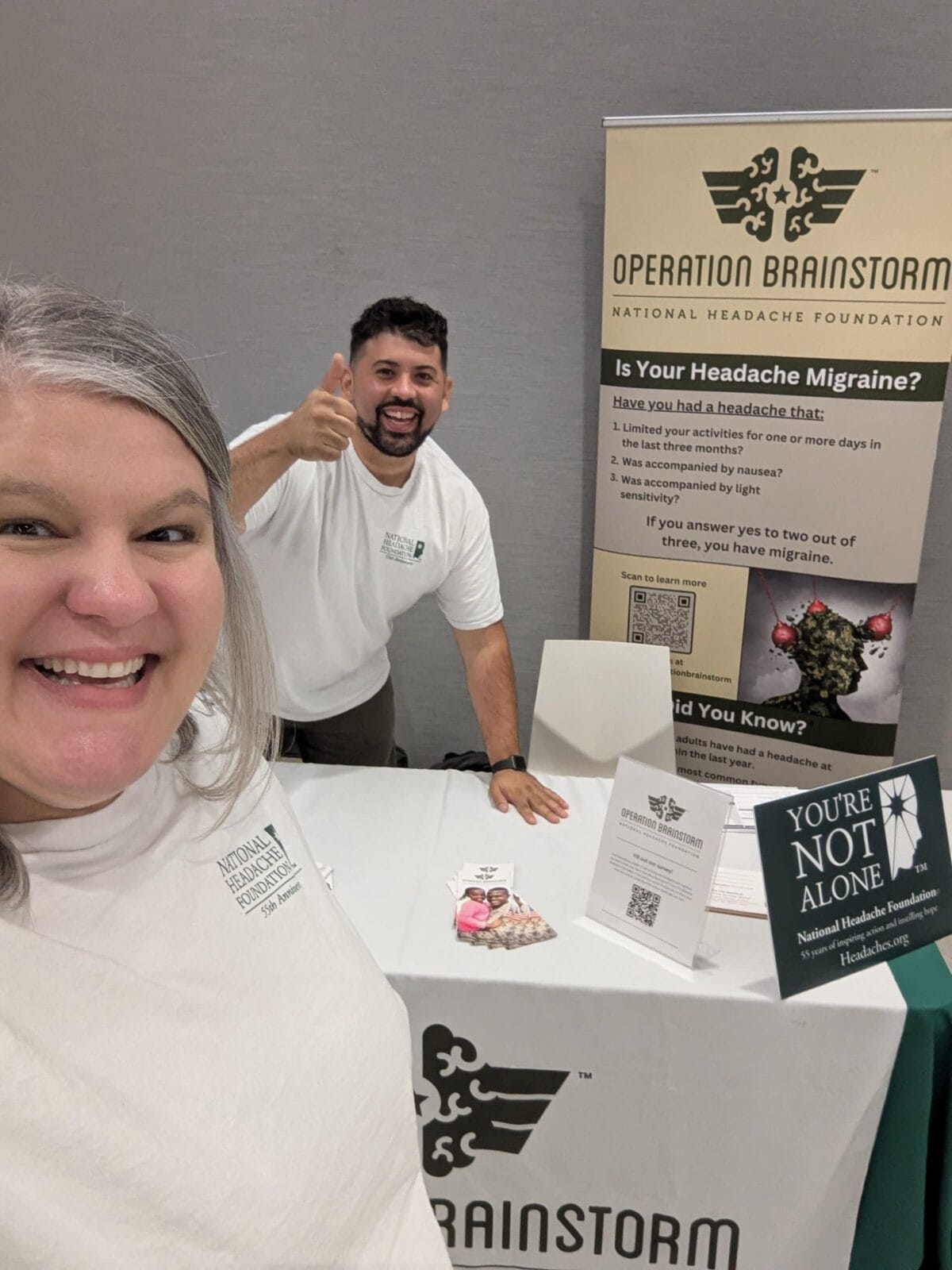 Two people at an informational booth for the National Headache Foundation's Operation Brainstorm, with pamphlets and signs about migraine awareness on the table.