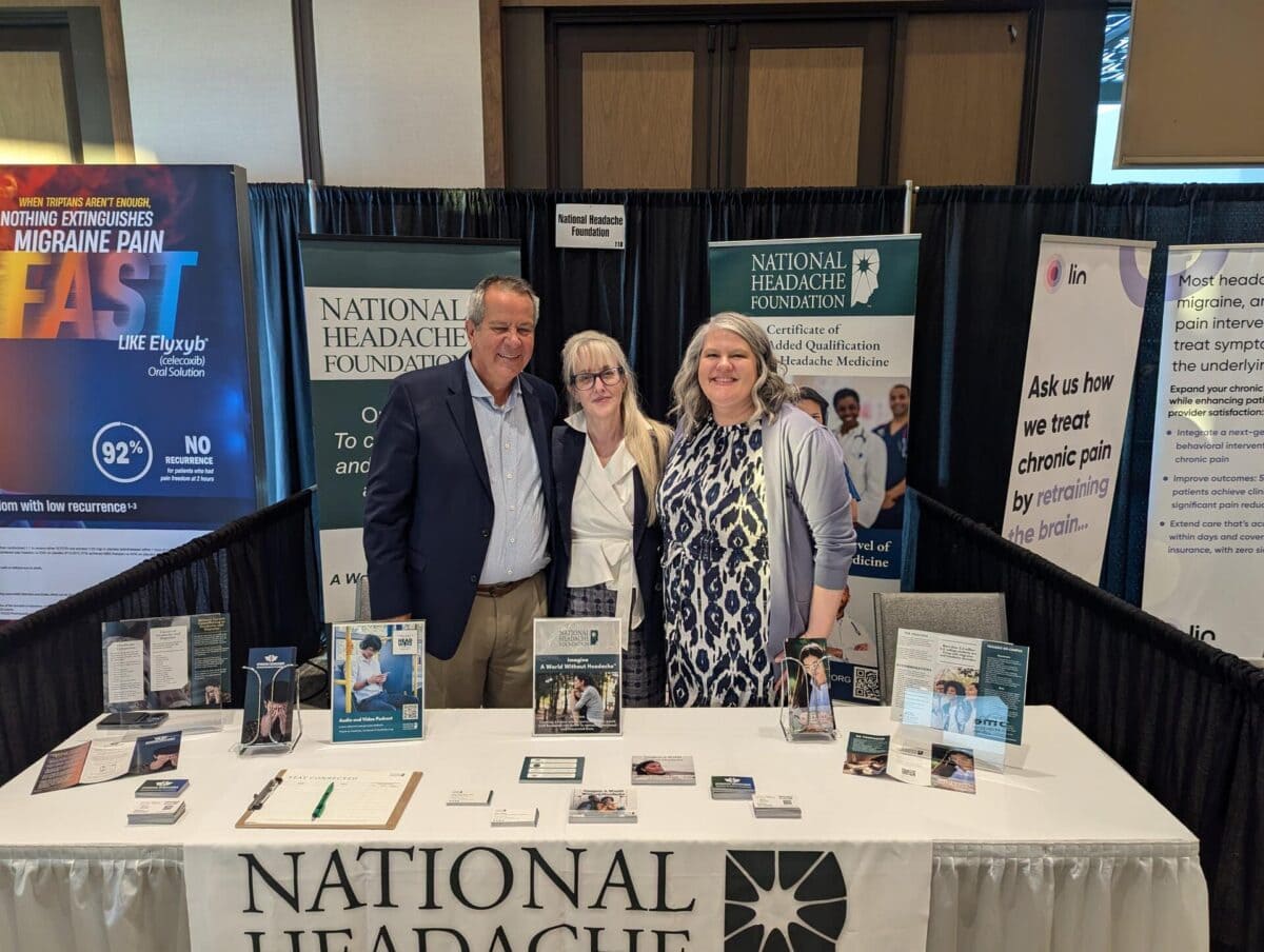 Three people stand behind a National Headache Foundation booth at an event, with informational materials and banners displayed on the table and in the background.