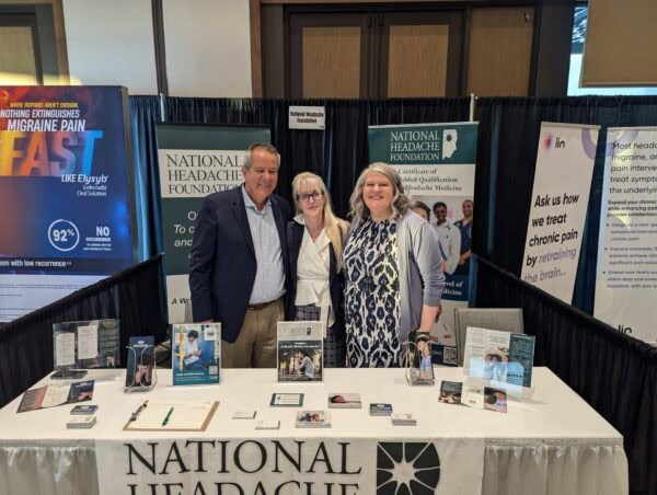 Three people stand behind a National Headache Foundation booth with informational materials and banners at a conference or event.
