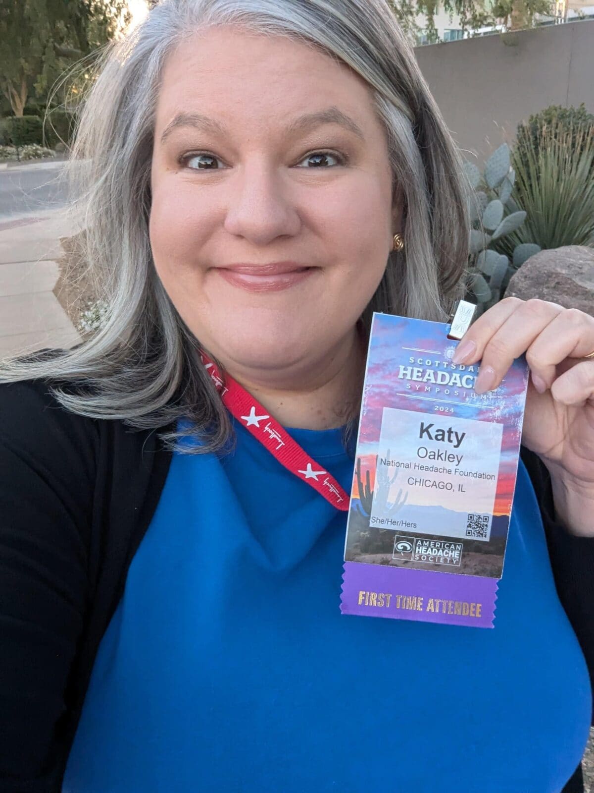 A woman with gray hair smiles at the camera while holding a conference badge that reads "Katy Oakley, FIRST TIME ATTENDEE." She is wearing a blue top and a red lanyard.