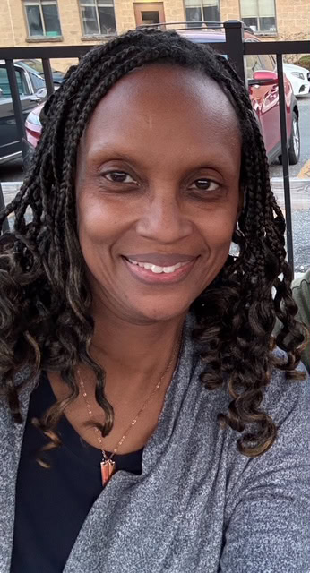 A woman with braided hair smiles at the camera while sitting outdoors, with parked cars and a building visible in the background.