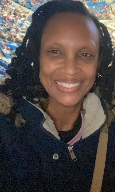 A woman wearing a dark coat with a fur-lined hood smiles at the camera in a crowded indoor arena.