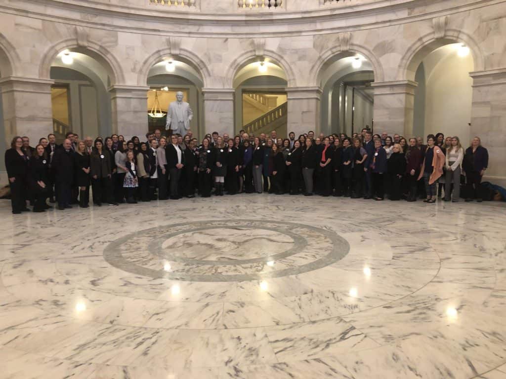 A large group of people stands together in a marble hall with arches, columns, and a statue in the background.