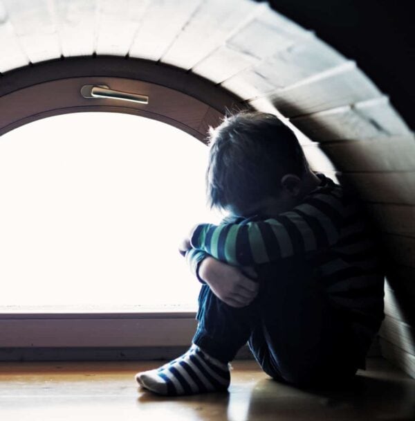 A child sits alone in a small arched alcove, hugging their knees, with light coming through a window behind them.