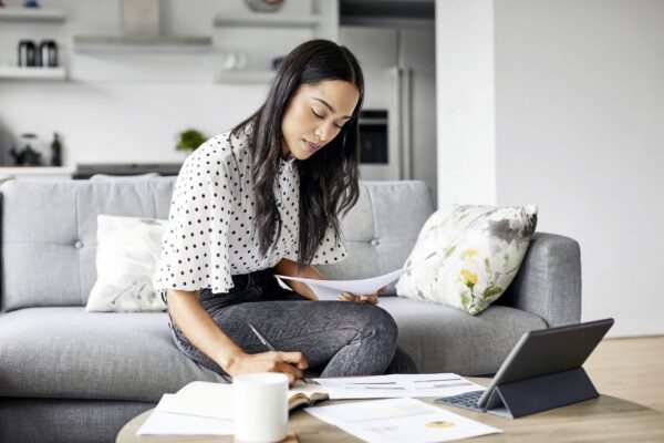 A woman sits on a couch working at a coffee table with a tablet, papers, and an open notebook in a modern living room.